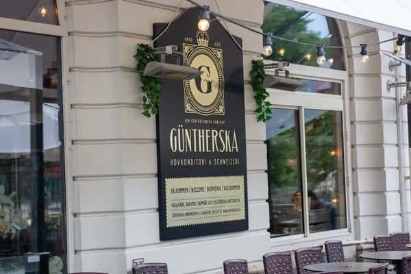 Svart skylt för "Güntherska Hovkonditori & Schweizeri" på vit fasad med fönster. Uteservering, värmare och ljusslingor