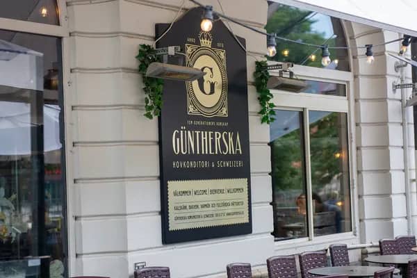 Svart skylt för "Güntherska Hovkonditori & Schweizeri" på vit fasad med fönster. Uteservering, värmare och ljusslingor