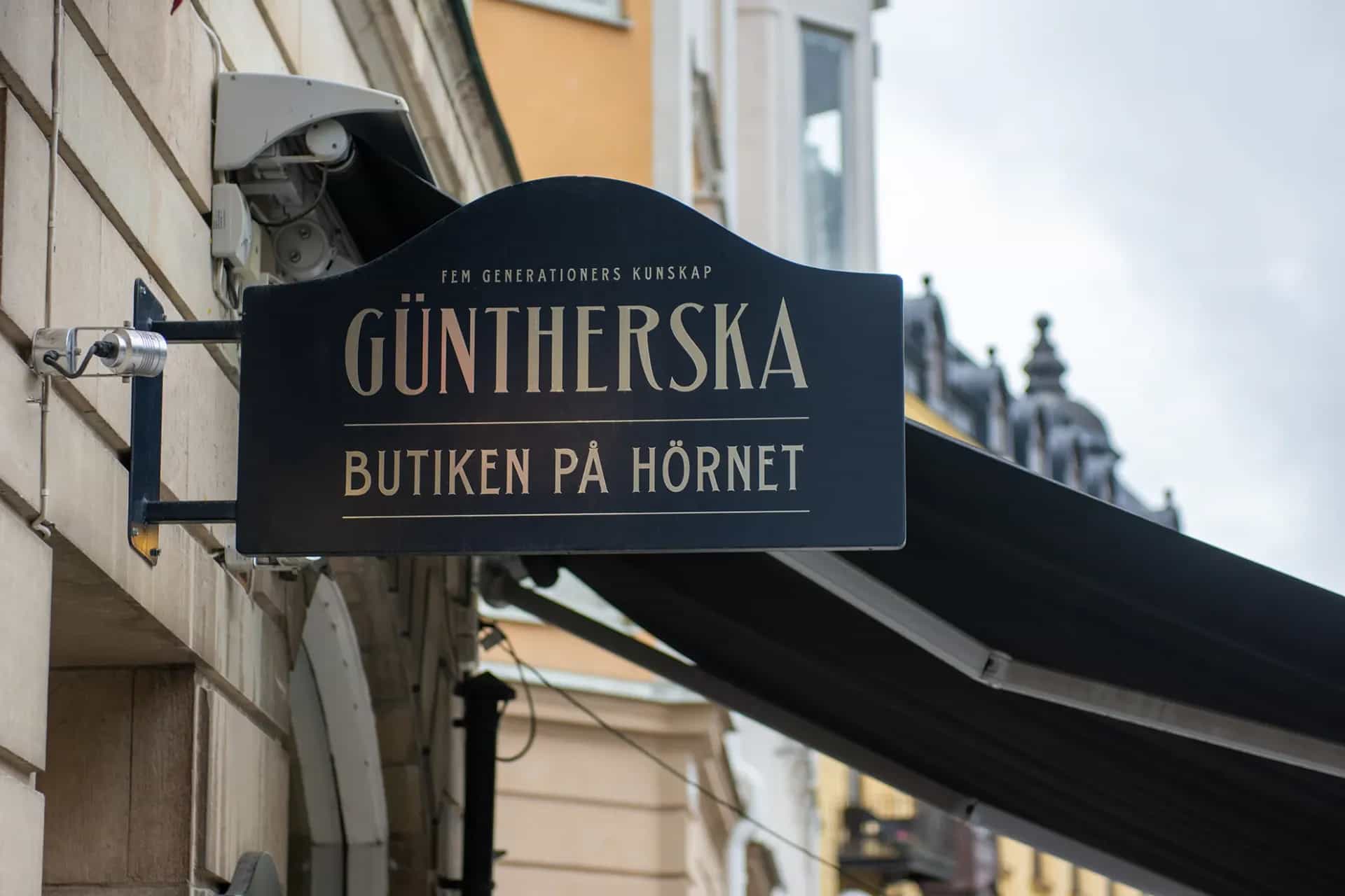 Svart butiksskylt för GÜNTHERSKA, med texten "Fem generationers kunskap" och "Butiken på hörnet", monterad på vägg