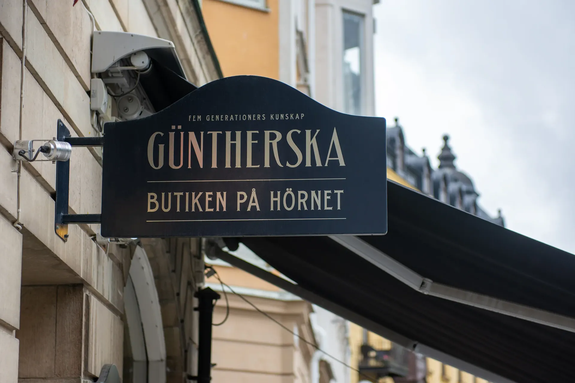 Svart butiksskylt för GÜNTHERSKA, med texten "Fem generationers kunskap" och "Butiken på hörnet", monterad på vägg