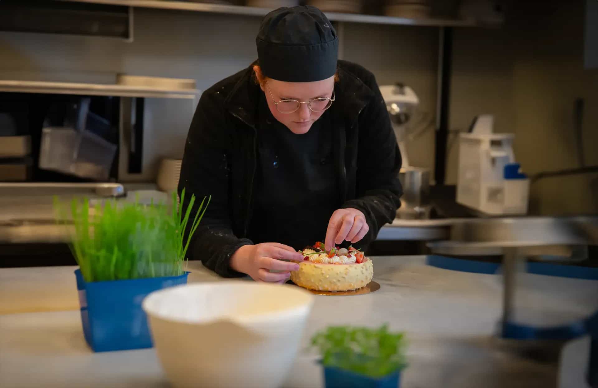 En person i kockmössa dekorerar en tårta med bär och grädde i ett kök. En kruka med gröna strån står till vänster