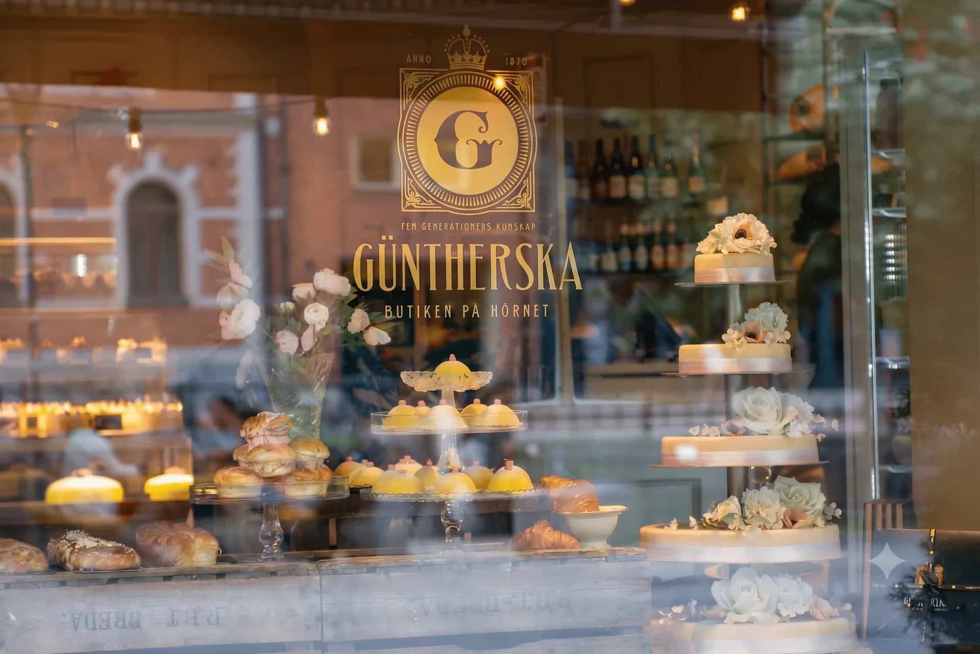 Tårtor, gula bakelser, bullar och bröd visas i Güntherska bageris skyltfönster. Logotyp och namn reflekteras i glaset